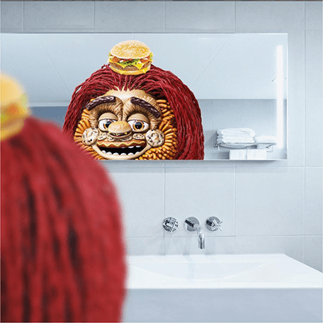 Image of Toomgis looking in the mirror of a bathroom. A hamburger sits on top of his head to symbolize a man bun.